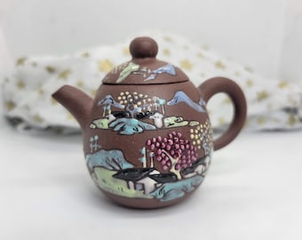 May include: A small brown teapot with a painted landscape design. The teapot has a lid and a handle. The design includes trees, mountains, and a river.