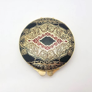 May include: A round, gold-toned compact mirror with a black and green enamel design. The design features intricate gold filigree and a red and gold diamond shape in the center.