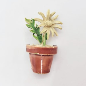 Puede incluir: Una flor blanca con hojas verdes en una maceta de cerámica marrón. La flor es una margarita con un centro amarillo y pétalos blancos. La maceta es un diseño simple con un fondo redondeado.