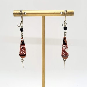 May include: A pair of dangle earrings with a terracotta-colored teardrop-shaped bead with a white design. The bead is attached to a silver wire with a black bead at the top.