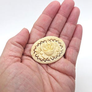 May include: Ivory-colored oval brooch with a carved rose design. The brooch has a scalloped edge and a detailed openwork border. The rose is the focal point, with delicate petals and a raised center. A vintage-inspired accessory.