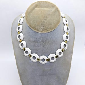 May include: A white, circular link necklace with gold-colored connectors. The necklace is displayed on a gray mannequin head. The links are oval-shaped with a circular opening in the center.