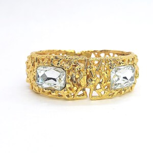 May include: A gold-tone hinged bracelet featuring a textured, branch-like metal surface and two large, rectangular, clear faceted crystals set on opposite sides of the band. The metal has a matte, organic finish with an intricate openwork design.
