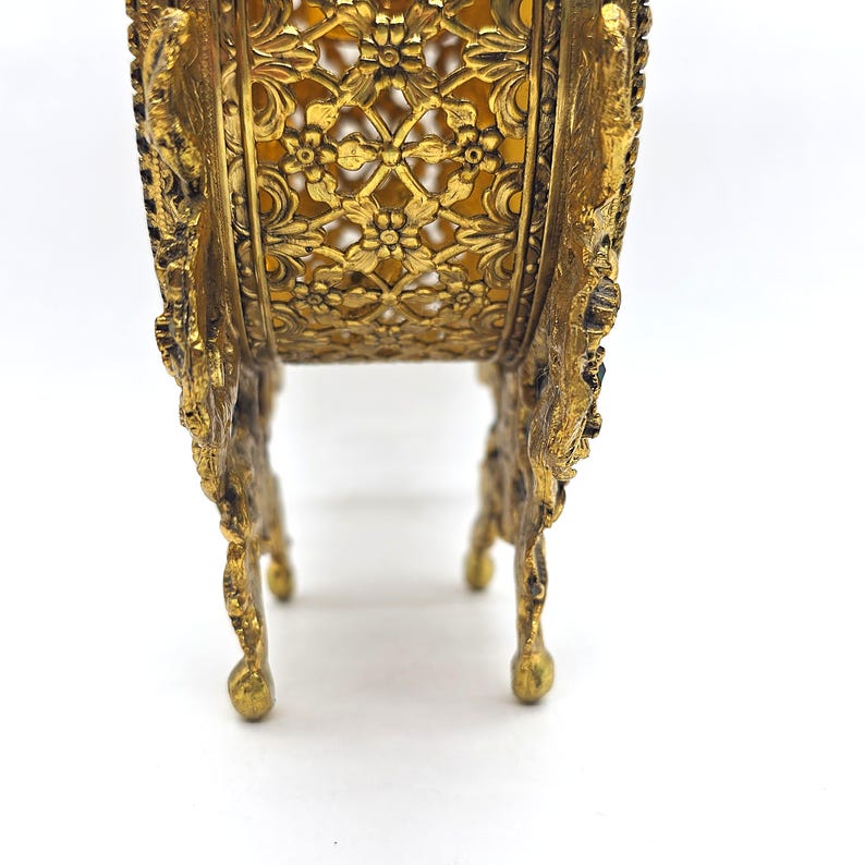 May include: Ornate gold-colored metal decorative stand with a filigree design. The stand has four legs and a detailed floral pattern around the sides. The interior appears to be a light yellow color.