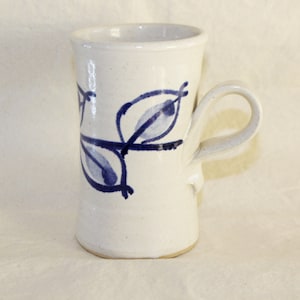 Puede incluir: Una taza de cerámica blanca con un diseño de hojas azul y blanco. La taza tiene un asa redondeada.