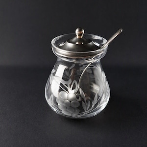 May include: A clear glass jar with a rounded body and a wide opening, featuring an etched floral pattern on the exterior. The jar includes a polished silver-toned metal lid with a central knob and a matching silver-toned metal spoon resting inside.