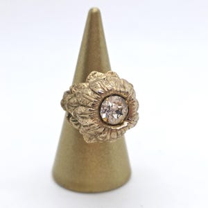 May include: A gold-toned ring with a large, clear gemstone at its center. The ring has a textured, floral-like design around the gemstone. The ring is displayed on a gold-colored cone-shaped ring holder against a white background.
