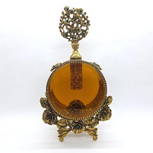 May include: An antique perfume bottle with a decorative gold-tone stand. The bottle has an amber-colored glass body and a detailed gold-tone stopper adorned with green gemstones. The stand features floral and leaf designs.