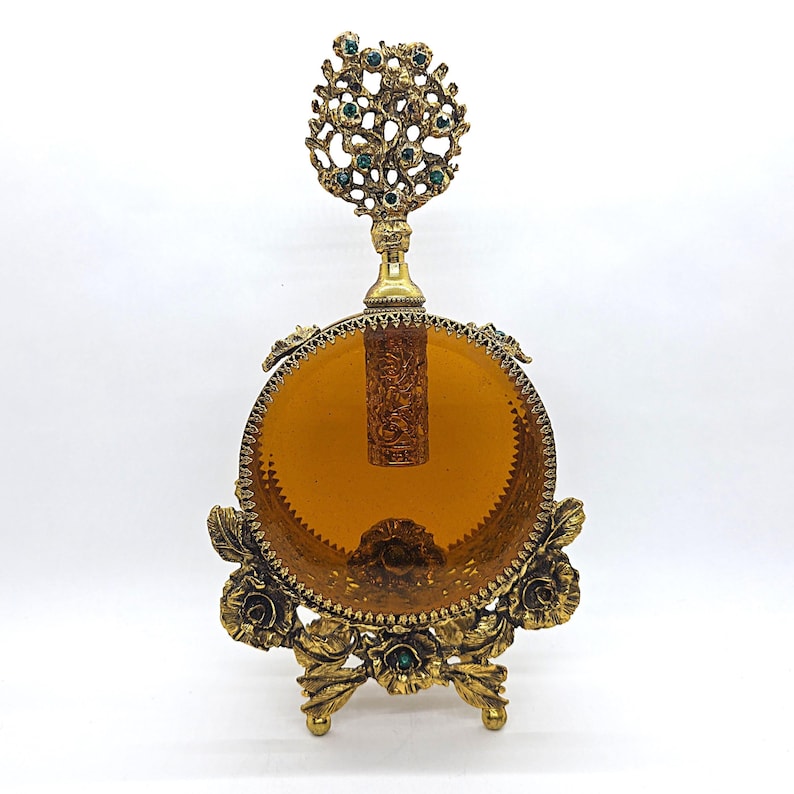 May include: An antique perfume bottle with a decorative gold-tone stand. The bottle has an amber-colored glass body and a detailed gold-tone stopper adorned with green gemstones. The stand features floral and leaf designs.