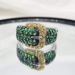 Pode incluir: Um anel de prata com um design de fivela de pedras verdes e amarelas.