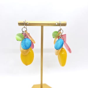 May include: A pair of dangle earrings with a mix of yellow, pink, blue, and green beads. The earrings are hanging from a gold stand.