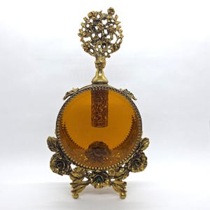 May include: An antique perfume bottle with a round amber-colored glass body, held in a decorative gold-toned metal frame. The frame features floral and leaf designs, with a tree-like stopper.