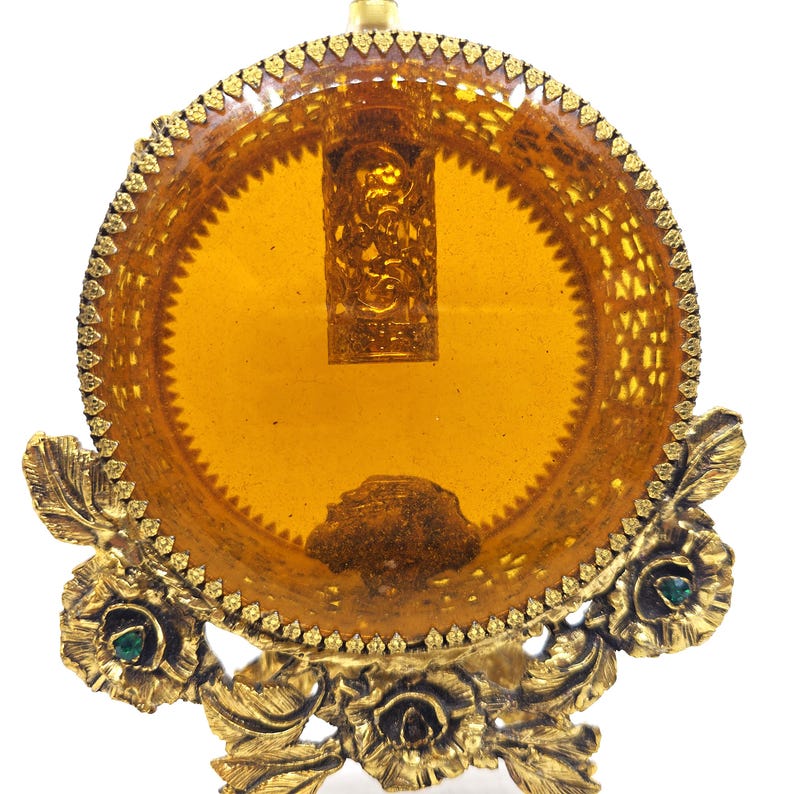 May include: An antique perfume bottle display. The bottle is amber-colored and encased in a gold-toned filigree holder. The base features floral and leaf designs with small green accents. The overall design is ornate and vintage.