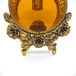May include: Ornate, antique gold-toned vanity tray with a round amber-colored glass insert. The tray features intricate floral and leaf detailing, with small green accents. The tray is supported by three decorative feet.