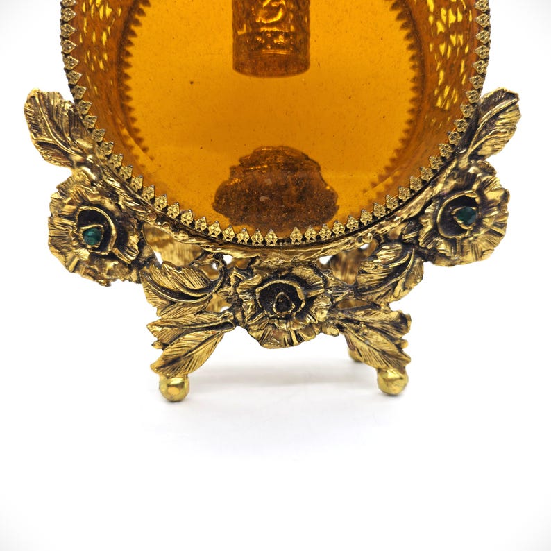 May include: Ornate, antique gold-toned vanity tray with a round amber-colored glass insert. The tray features intricate floral and leaf detailing, with small green accents. The tray is supported by three decorative feet.