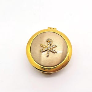May include: A gold-toned compact mirror with a circular design. The mirror features a gold border and a central emblem adorned with sparkling rhinestones. The emblem is a stylized symbol, possibly religious or symbolic.