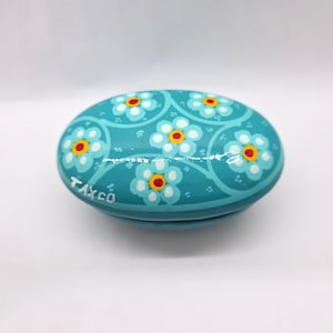 May include: Oval-shaped, turquoise trinket box with a floral design. The box features white flowers with yellow centers and red dots, arranged in a repeating pattern. The text "TAXCO" is visible on the side of the box.