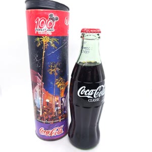 May include: A Coca-Cola Classic bottle with a red cap next to a cylindrical container. The container is red at the top with "100 Years of Magic" and a Mickey Mouse graphic. The container has a nighttime scene with palm trees and buildings.