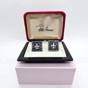 Puede incluir: Un par de gemelos cuadrados con diseño de flor de lis, presentados en una caja forrada de terciopelo negro y rojo. Los gemelos son negros con borde blanco y un acento rojo. La caja está etiquetada como "Styled by Fifth Avenue".