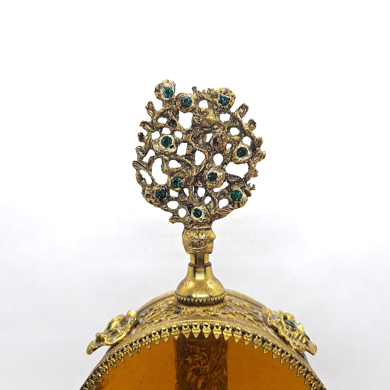 May include: A vintage perfume bottle with a gold-colored filigree design. The bottle's top features a decorative tree-like ornament adorned with green gemstones. The bottle has an amber-colored glass base and ornate detailing.