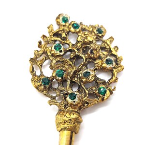 May include: A gold-toned key with a floral design, adorned with emerald-green gemstones. The key's head features an intricate, openwork pattern of leaves and flowers. The key's stem is a simple, smooth gold color.