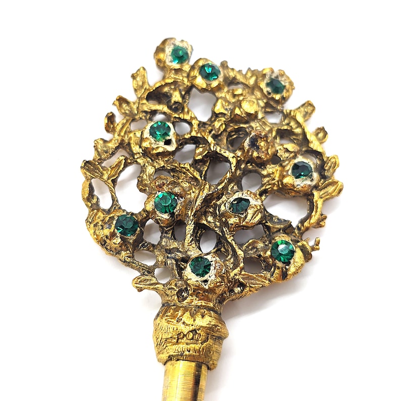 May include: A gold-toned key with a floral design, adorned with emerald-green gemstones. The key's head features an intricate, openwork pattern of leaves and flowers. The key's stem is a simple, smooth gold color.