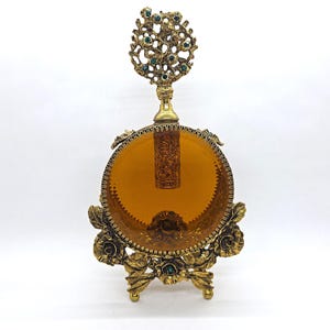 May include: An antique perfume bottle with a round amber glass body and ornate gold-tone metalwork. The bottle features a decorative floral base and a detailed top with green accents. The stopper is visible inside the bottle.
