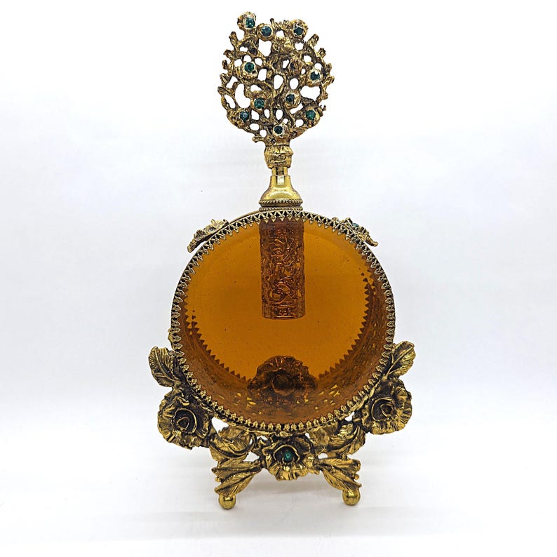 May include: An antique perfume bottle with a round amber glass body and ornate gold-tone metalwork. The bottle features a decorative floral base and a detailed top with green accents. The stopper is visible inside the bottle.