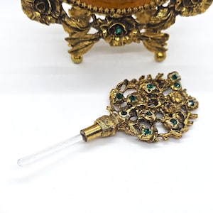 May include: Ornate gold-toned perfume bottle stopper with a clear glass applicator. The stopper features a floral design with embedded green gemstones. The background shows a matching perfume bottle base with floral detailing.