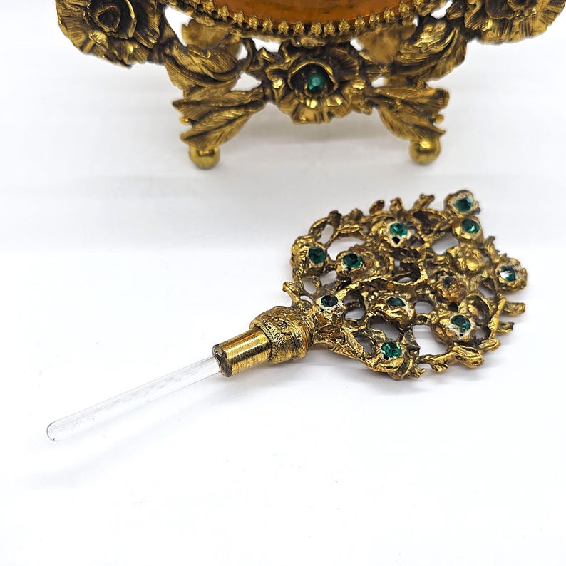 May include: Ornate gold-toned perfume bottle stopper with a clear glass applicator. The stopper features a floral design with embedded green gemstones. The background shows a matching perfume bottle base with floral detailing.