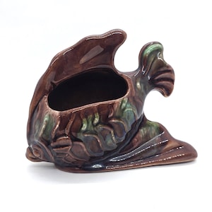 Puede incluir: Una maceta de cerámica con forma de pez en tonos marrones, verdes y azules. La maceta tiene una gran abertura para plantas y un diseño texturizado en forma de escamas. El pez tiene una cola curvada y una aleta que se extiende hacia arriba.