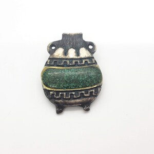 Op de afbeelding: Een zilveren hanger in de vorm van een klein, groen en goud gedecoreerd potje met een geometrisch ontwerp.