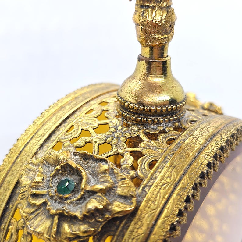 May include: Close-up of a vintage perfume bottle with ornate gold filigree and a green gemstone accent. The bottle features a detailed floral design and a decorative stopper, showcasing an antique aesthetic.