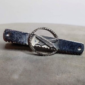 May include: Silver tie clip with a saw blade design and a circular, detailed design in the center.