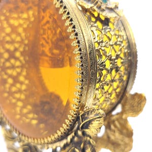 May include: Close-up of a vintage, ornate, gold-colored decorative object. The object features a transparent amber-colored center, intricate filigree, and floral accents. The design includes a detailed, sculpted figure at the base.
