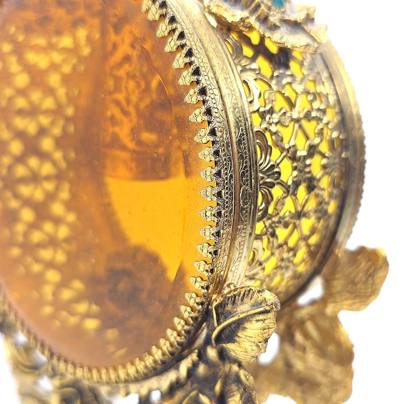 May include: Close-up of a vintage, ornate, gold-colored decorative object. The object features a transparent amber-colored center, intricate filigree, and floral accents. The design includes a detailed, sculpted figure at the base.