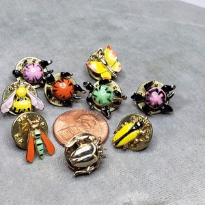 May include: Assortment of nine colorful enamel insect pins. The pins feature a variety of insects, including a bee, a butterfly, a ladybug, and a beetle. The pins are all gold-toned and have a black backing.