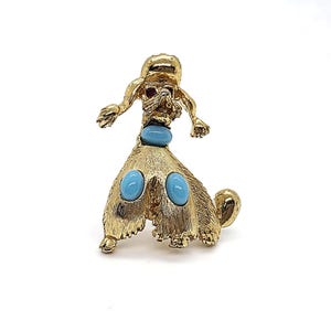 May include: A gold-tone poodle brooch featuring a textured metal finish. The dog figure is adorned with three smooth, oval-shaped light blue cabochon stones set on the chest and legs, and a single small red stone serving as the eye of the poodle.