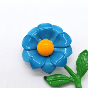 May include: A vintage flower brooch featuring a bright blue flower with an orange center. The brooch has a green stem and leaf. This floral accessory is a charming addition to any outfit.