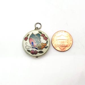Puede incluir: Un colgante redondo de esmalte blanco con un diseño de mariposa colorido. La mariposa tiene alas azules, rosas, verdes y amarillas. El colgante está rodeado de pequeñas flores rojas. El colgante es aproximadamente del tamaño de una moneda de un céntimo de euro.