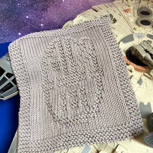 May include: A gray knitted dishcloth with a textured pattern. The dishcloth is lying on top of a model of the Millennium Falcon spaceship.