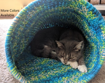 Peut inclure: Un chat gris et blanc se repose dans un lit pour chat crocheté. Le lit est fait de fil bleu, vert et blanc. Le texte "Plus de couleurs disponibles" est visible dans le coin supérieur gauche de l'image.