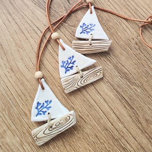 Puede incluir: Tres colgantes de cerámica con forma de velero con cordones de cuero marrón. Las velas blancas presentan diseños de coral azul. Las bases de los barcos de madera tienen un patrón de vetas de madera natural.