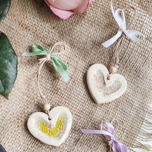 May include: Heart-shaped ceramic ornaments with a textured surface and a small hole for hanging. Each heart is cream-colored and adorned with a ribbon in various colors, including green, white, and purple. A small wooden bead is attached to each ornament.