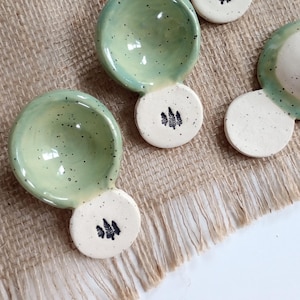 May include: Handmade ceramic spoon rests with a light green glaze and speckled finish. Each rest features a small, round base with a black tree design. The spoon rests are arranged on a burlap surface.