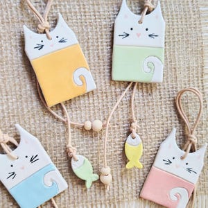 Op de afbeelding: Vier keramische katvormige ornamenten met visbedels. Elke kat heeft een wit gezicht met zwarte snorharen en een roze neus. De ornamenten zijn geel, groen, blauw en roze. De ornamenten hangen aan een touwtje.