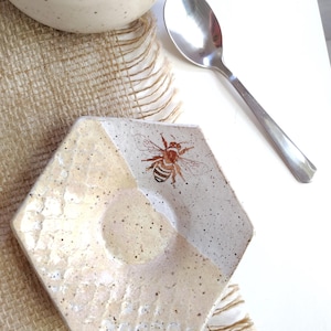 May include: A hexagonal ceramic dish with a bee illustration. The dish is off-white with speckled detailing and a recessed center. A stainless steel spoon and a speckled ceramic mug are also visible.