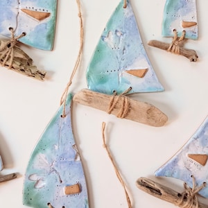 Könnte beinhalten: Handgefertigte Keramik-Segelboot-Ornamente mit blau-türkisfarbenen Segeln, befestigt an Treibholzsockeln mit Bindfaden. Jedes Segel hat einen kleinen, rechteckigen braunen Akzent. Natürliche Bindfaden-Schlaufen sind zum Aufhängen angebracht.
