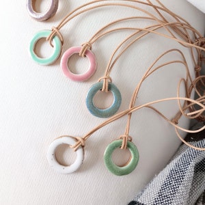 Puede incluir: Una colección de collares con colgantes de cerámica hechos a mano. Cada collar presenta un colgante circular en diferentes colores, incluyendo rosa, verde, azul, blanco y morado. Los colgantes están ensartados en cordones de cuero color canela.