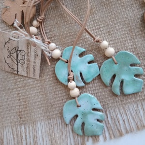 May include: Monstera leaf-shaped ceramic essential oil diffusers. The light blue leaves have a speckled design and are strung on leather cords with wooden beads. Tags read "Ceramic & Leather Essential Oil Diffuser."